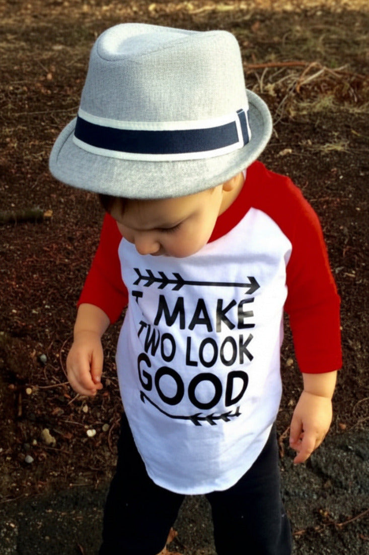 A toddler wearing a light gray fedora hat with a navy band and a white and red raglan shirt that says 'MAKE TWO LOOK GOOD' in black text, standing outdoors on a dirt ground.